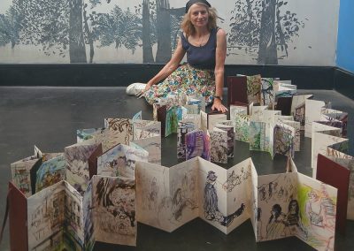 The artist is sitting on the floor surrounded by concertina sketchbooks.